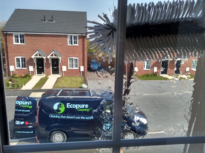 A view looking out a window. There is a blue van outside with "Ecopure Cleaning" written on the side and a window cleaning brush with water scrubbing the glass. Across the road you can see red brick houses with black doors.